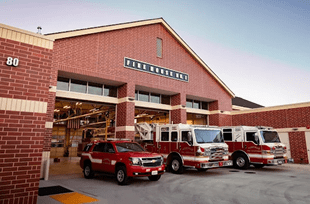 Red brick fire station exterior featuring two fire trucks and an emergency vehicle parked outside. The image highlights the facility's role in public safety.