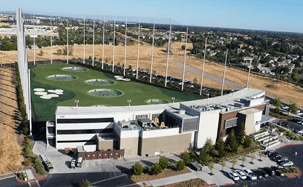 Aerial view of a modern golf driving range featuring multiple hitting bays, green turf, and tall netting, surrounded by open land and parking.