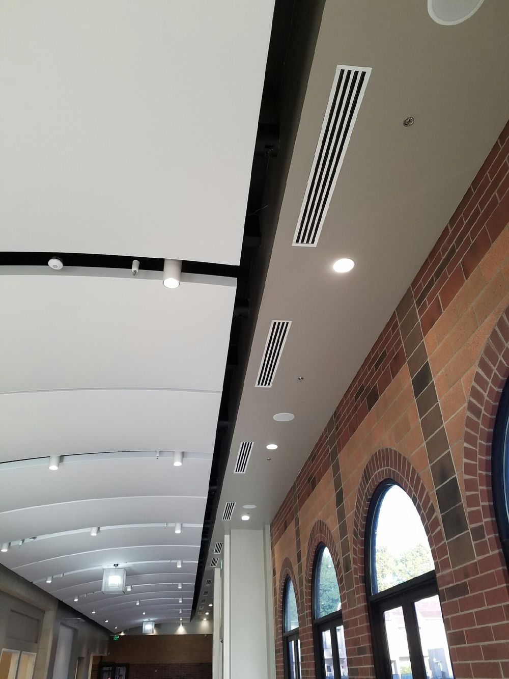 Modern corridor featuring an arched ceiling with recessed lighting and air vents, alongside a brick wall with large arched windows.